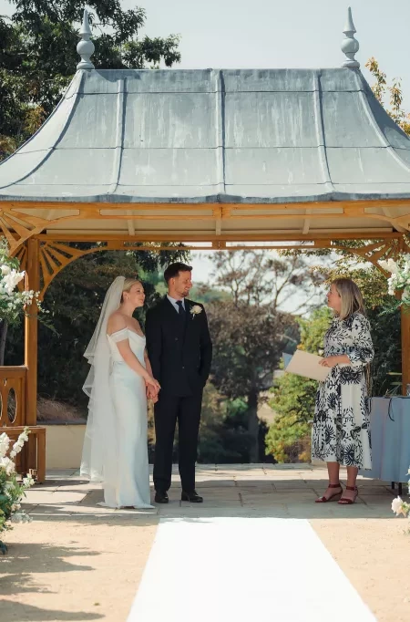 A bride and groom standing with their celebrant - Sam White - underneath the pagoda at Clevedon Hall.