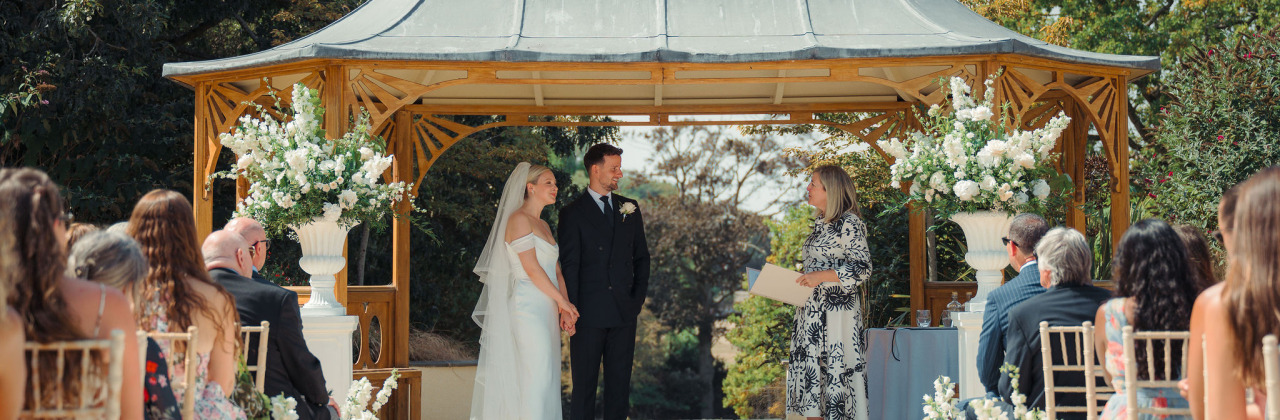 A bride and groom standing with their celebrant - Sam White - underneath the pagoda at Clevedon Hall.